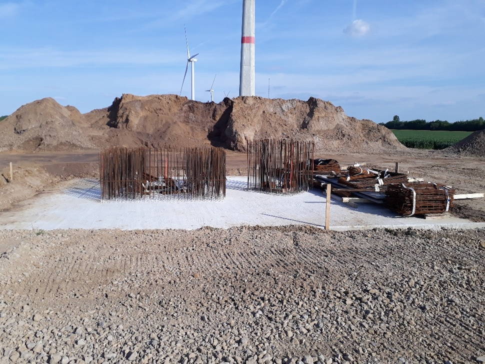 The crane foundation is larger than the pedestals, reinforcements protrude from the concrete, right four heaps with further reinforcements are stacked on the concrete, the earth around the foundation is rolled flat.